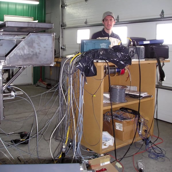 Mathieu steht vor seinem Computer. Auf einem hohen Tisch, von dem auf beiden Seiten zahlreiche Kabel herabhängen.  