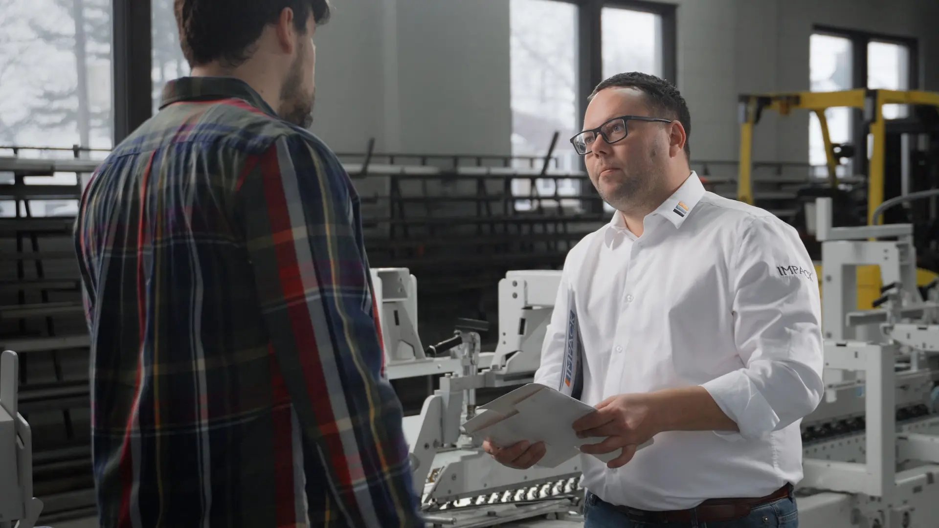 Two men discuss carton packaging near a folder-gluer