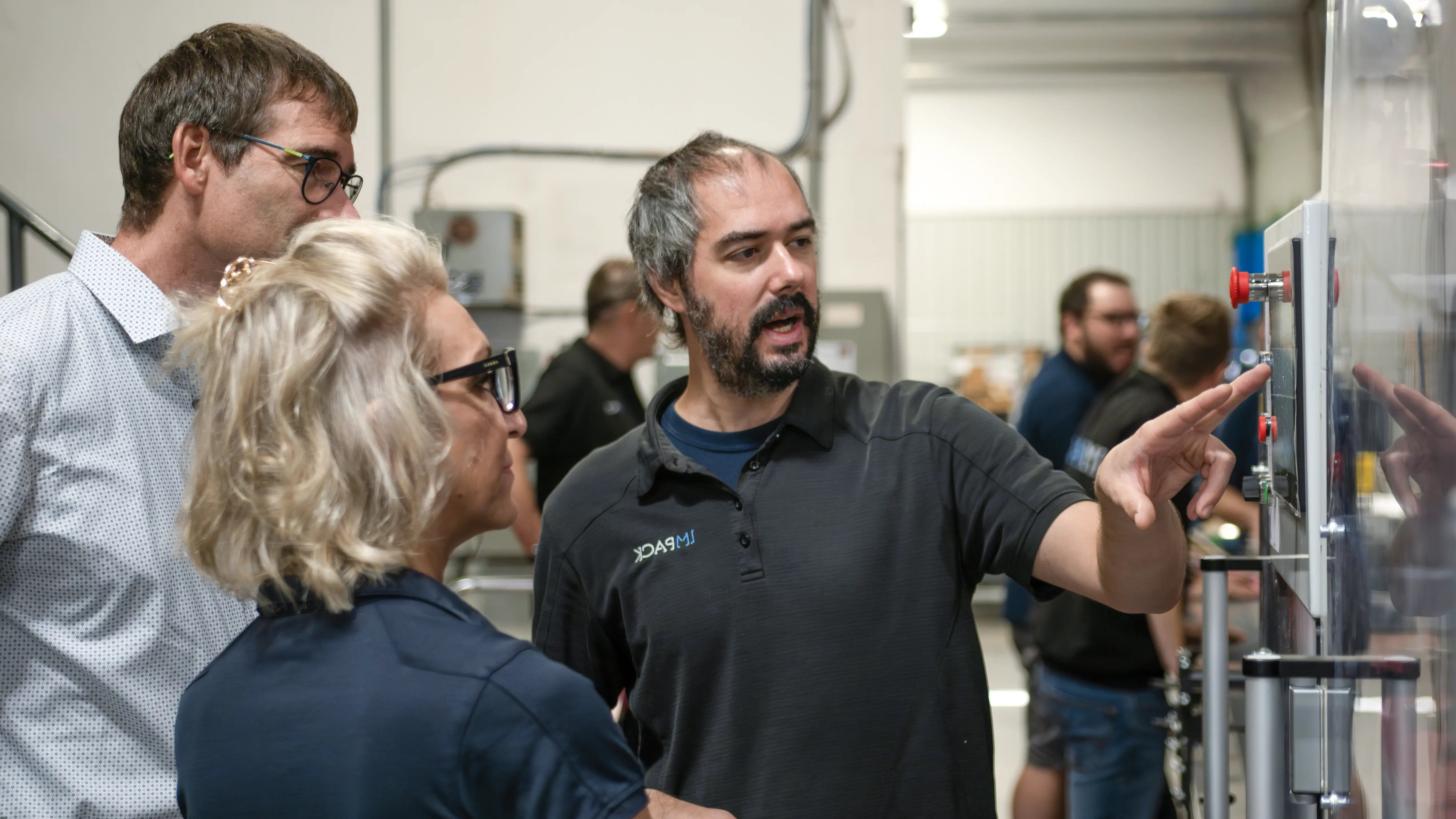 Jonathan, IMPACK’s service coordinator, a male client and Natascha, IMPACK’s Sales Director for german speaking countries, stand next to the MFA. Jonathan gestures toward the HMI screen.